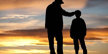 A silhouette of a father and son sharing a tender moment. Additional themes include single parent, parenting, father, fatherhood, stepfather, consoling, care, unity, family, bonding, encouragement, coach, role model, instructor, guidance, and comforting.